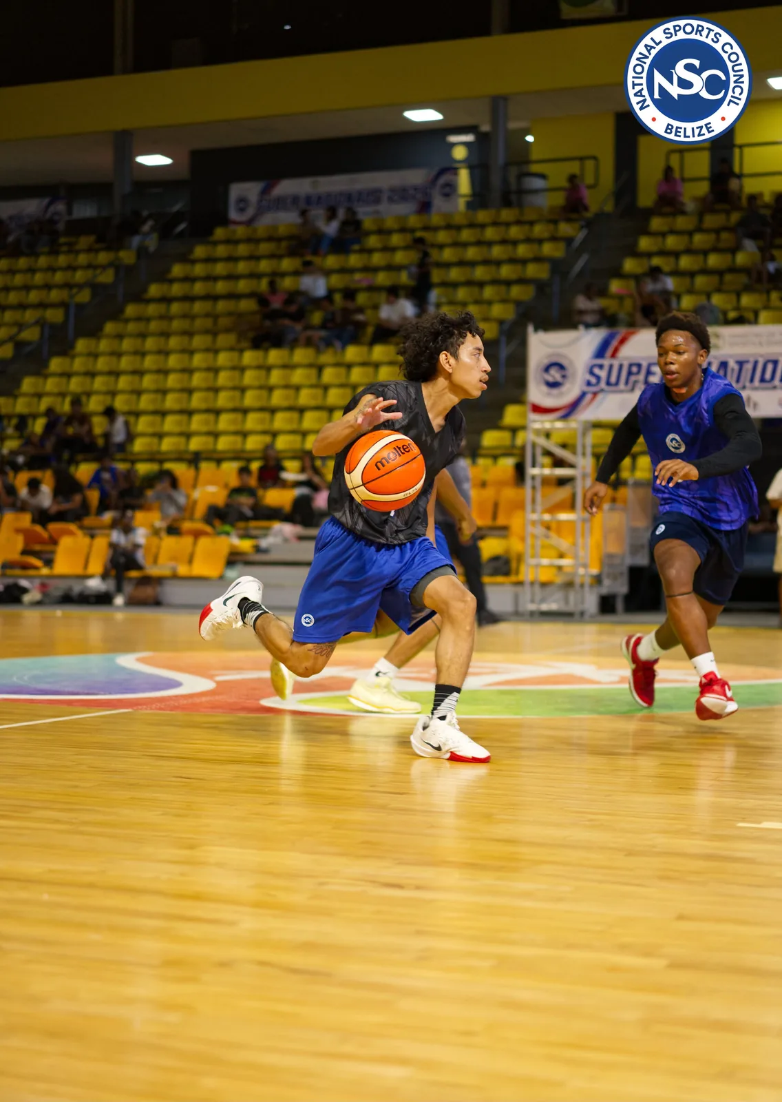 A basketball player driving toward the basket past a defender, with the Super Nationals 2026 banner behind them